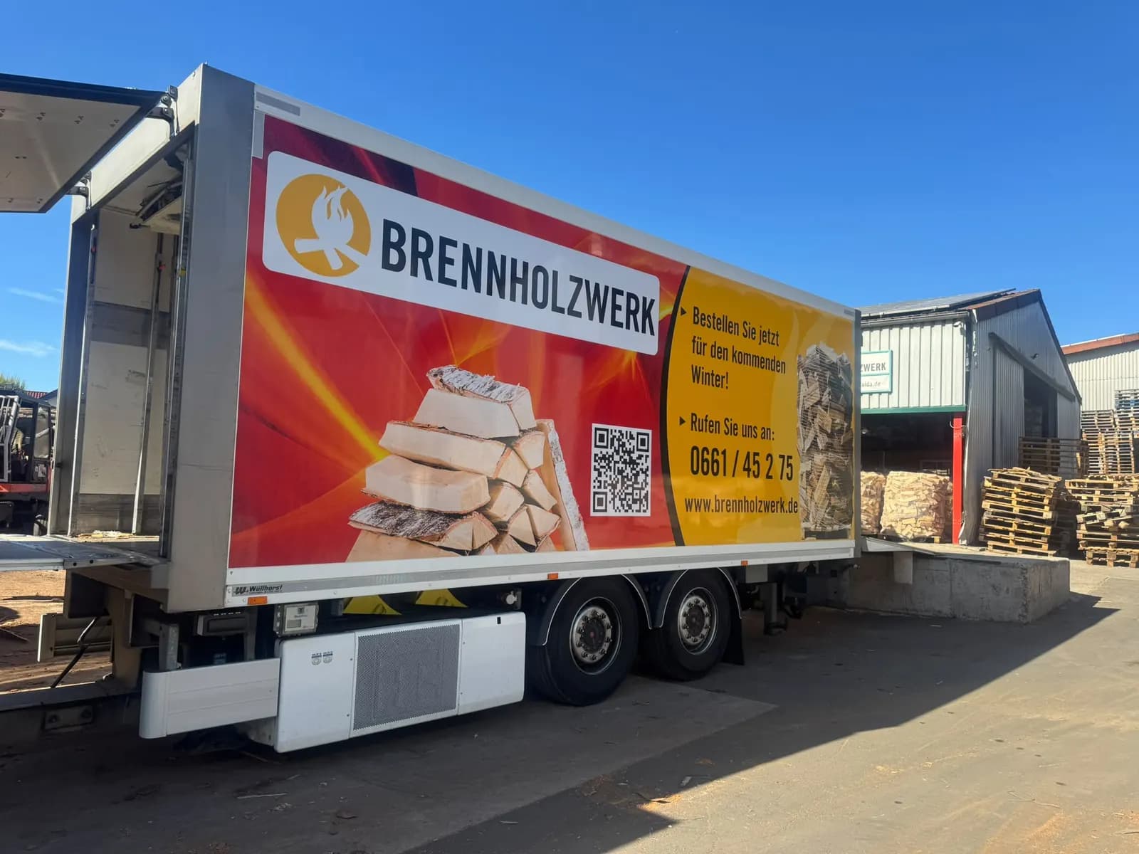 Brennholzwerk-Anhänger an der Laderampe, daneben verpackte Brennholzpaletten